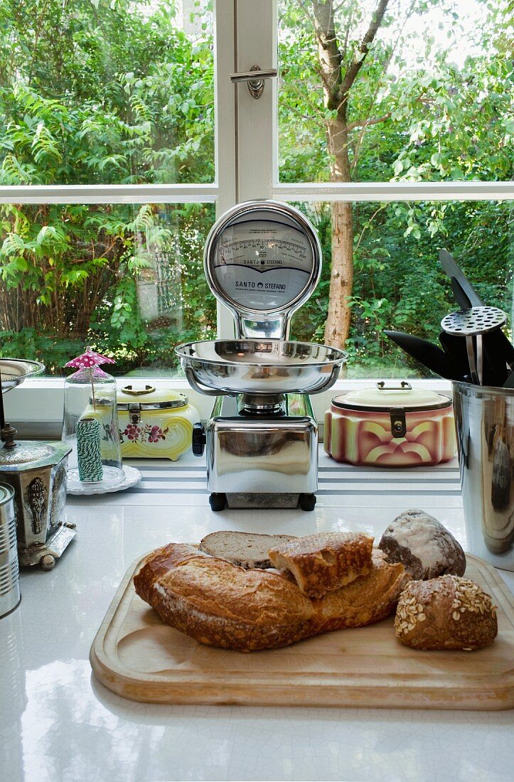 Bread on chopping board and various kitchen utensils on ceramic worksurface in front of kitchen window