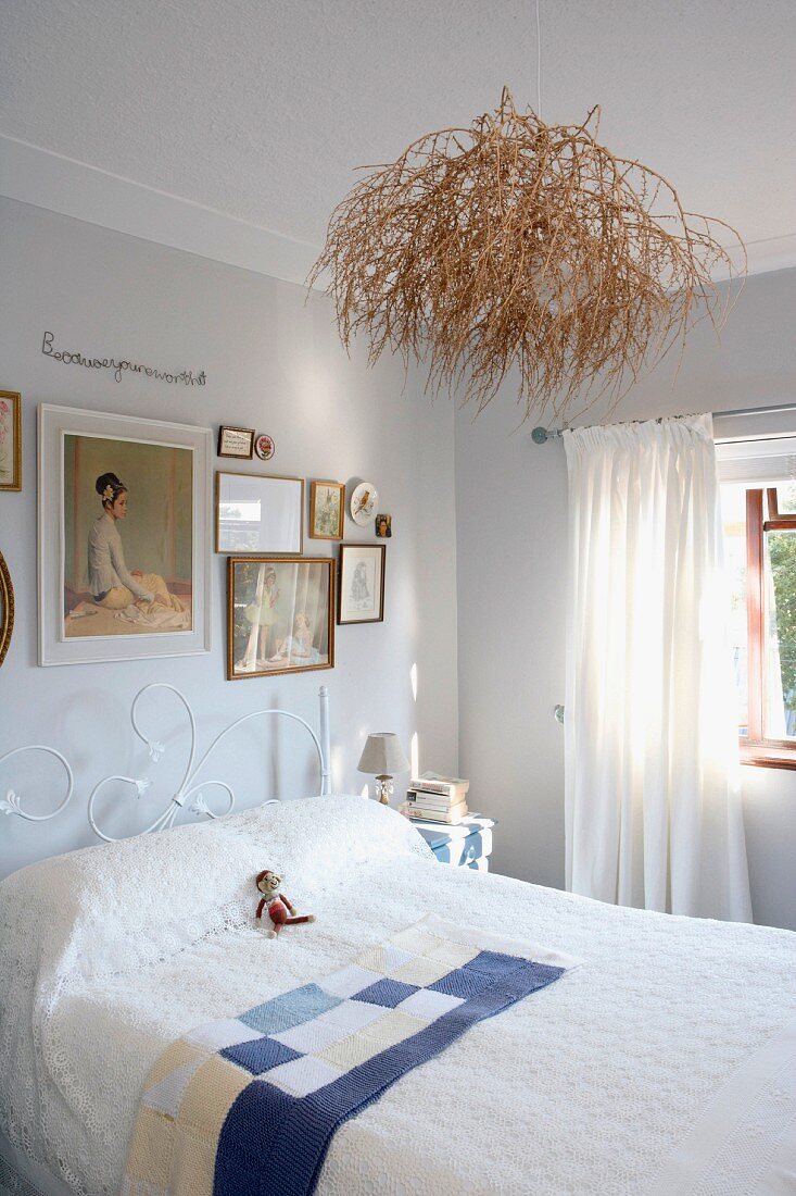 Bright bedroom with bundle of twigs on ceiling lamp above bed with white, delicate metal frame below framed pictures on wall