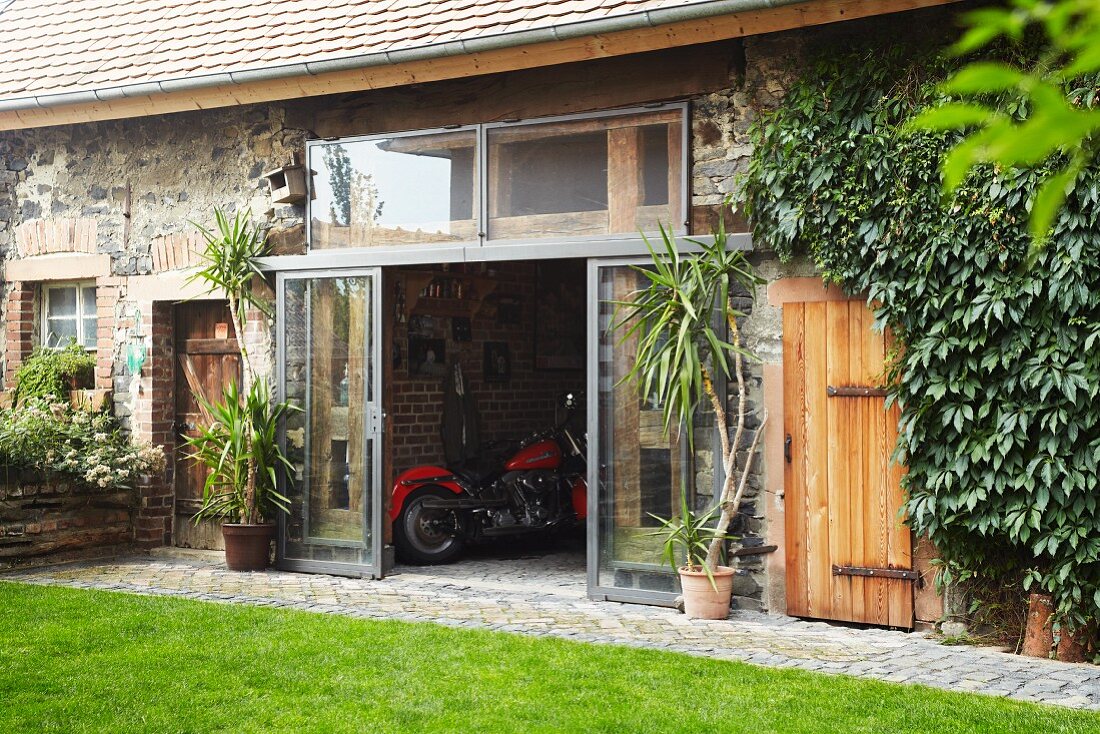 Landhaus mit renoviertem Schuppen und Blick durch offene Glas Schiebetür auf rotes Motorrad