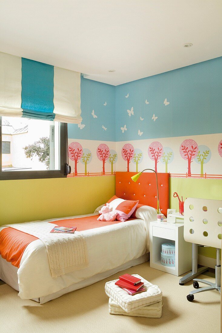 Bett mit Kopfpolster vor farbenfroher Wandgestaltung in Schablonentechnik mit stilisiertem Landschaftsmotiv