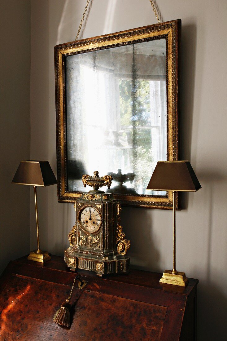 Antike Uhr von Lampen umrahmt auf einem Sekretär und Goldrahmen-Spiegel an der Wand