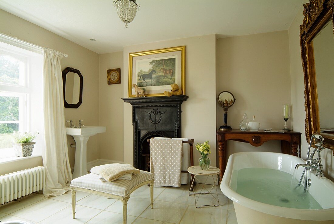 Badezimmer mit offenem Kamin und weißem Fliesenboden, über der gefüllten Badewanne ein großer Goldrahmenspiegel, davor eine Polsterbank mit Handtüchern und im Hintergrund ein Waschtisch