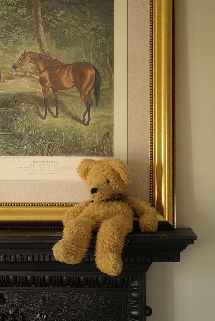 Beiger Teddybär sitzt locker auf dunklem Kaminsims vor angelehntem Bild mit Goldrahmen, es zeigt ein gezeichnetes Pferd in der Natur