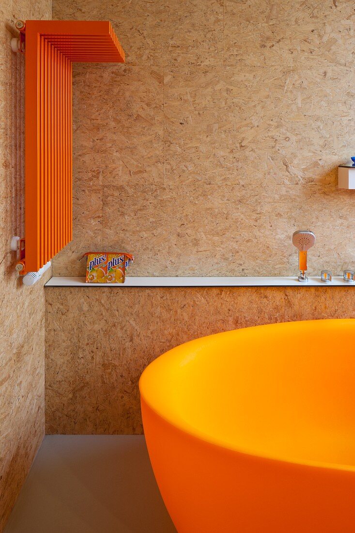 Badezimmerecke mit Handtuchtrockner in Orange an Spanplatten-Wand und teilweise sichtbarer Badewanne