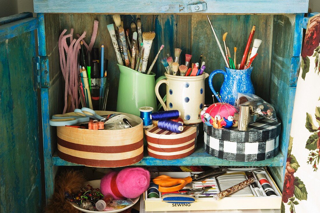 Blue wooden cupboard with paintbrushes, pens and sewing materials in patterned boxes and jugs
