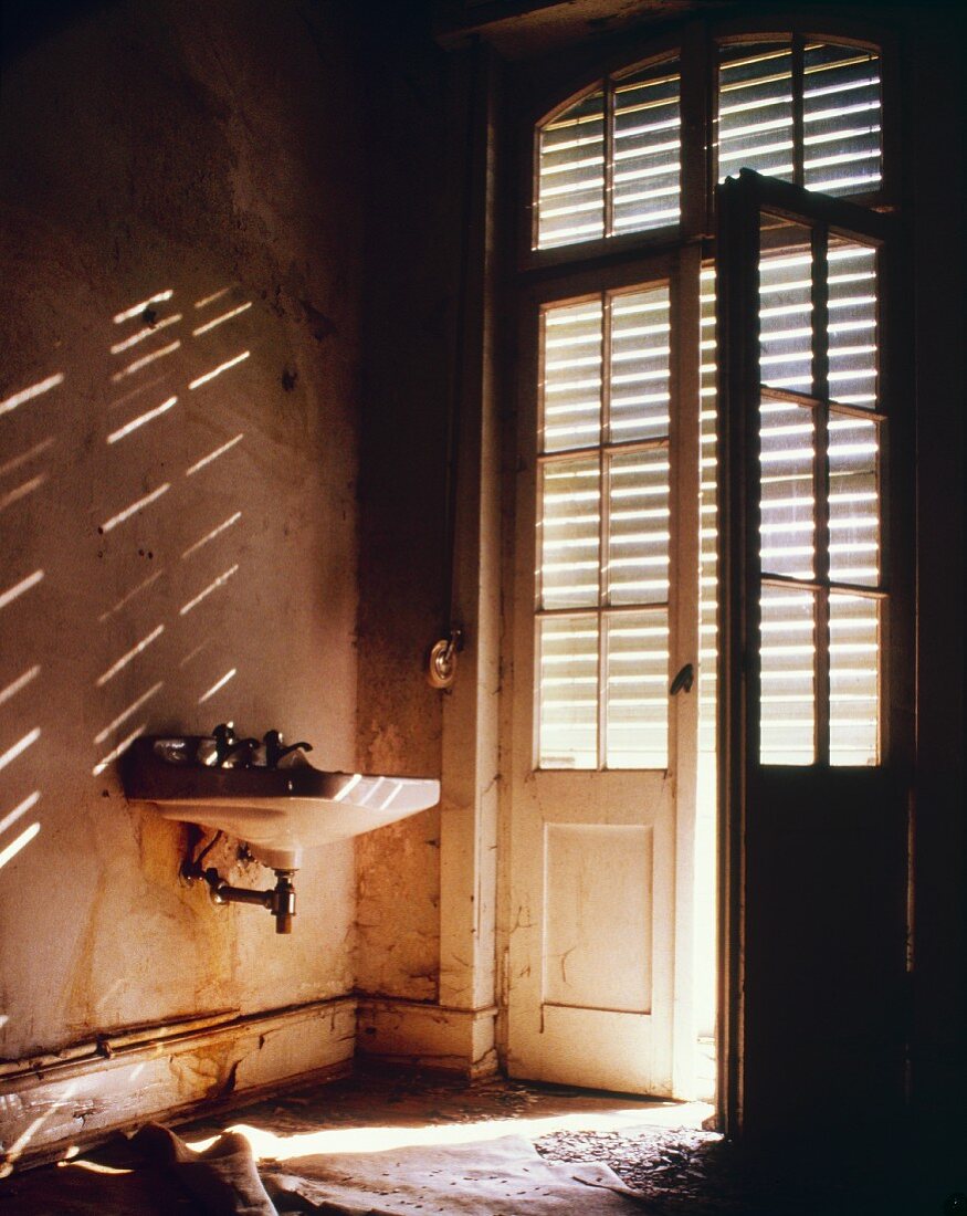 Vintage Waschbecken neben Tür mit Glaseinsatz und Jalousie in einen verlassenem Haus