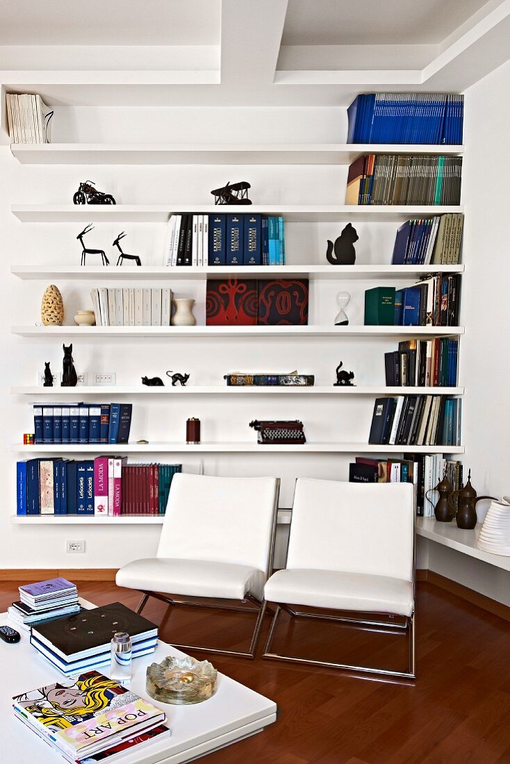 Two white, designer armchairs in front of book sets and ornaments on white, minimalist shelves