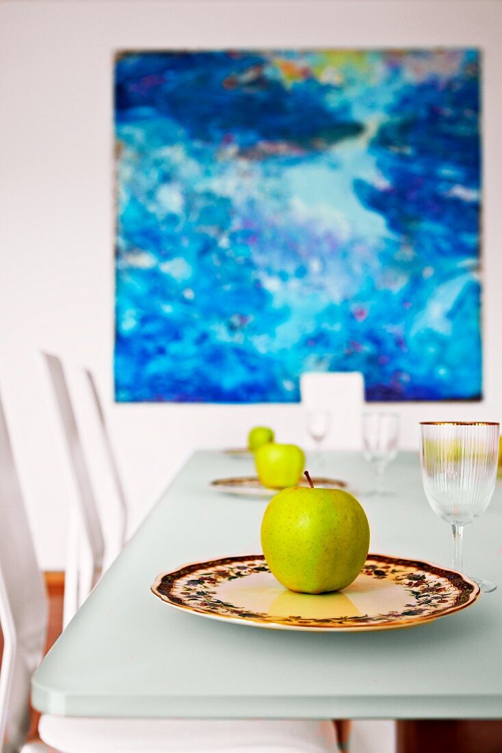 Green apples on decorative china plates in front of oil painting in shades of blue