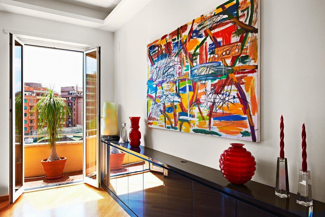 Abstract painting above glossy black sideboard and open balcony door with view of city