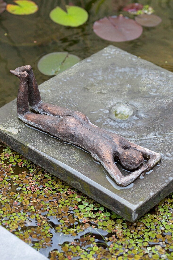 Liegende Bronzefigur auf Bronzefloß zwischen Wasserlinsen und Seerosen
