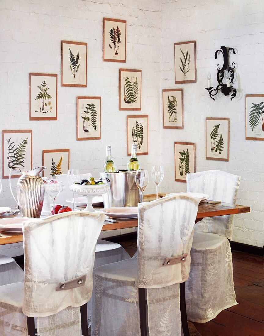 Covered chairs at set dining table in corner of room with framed pictures of ferns on whitewashed brick walls