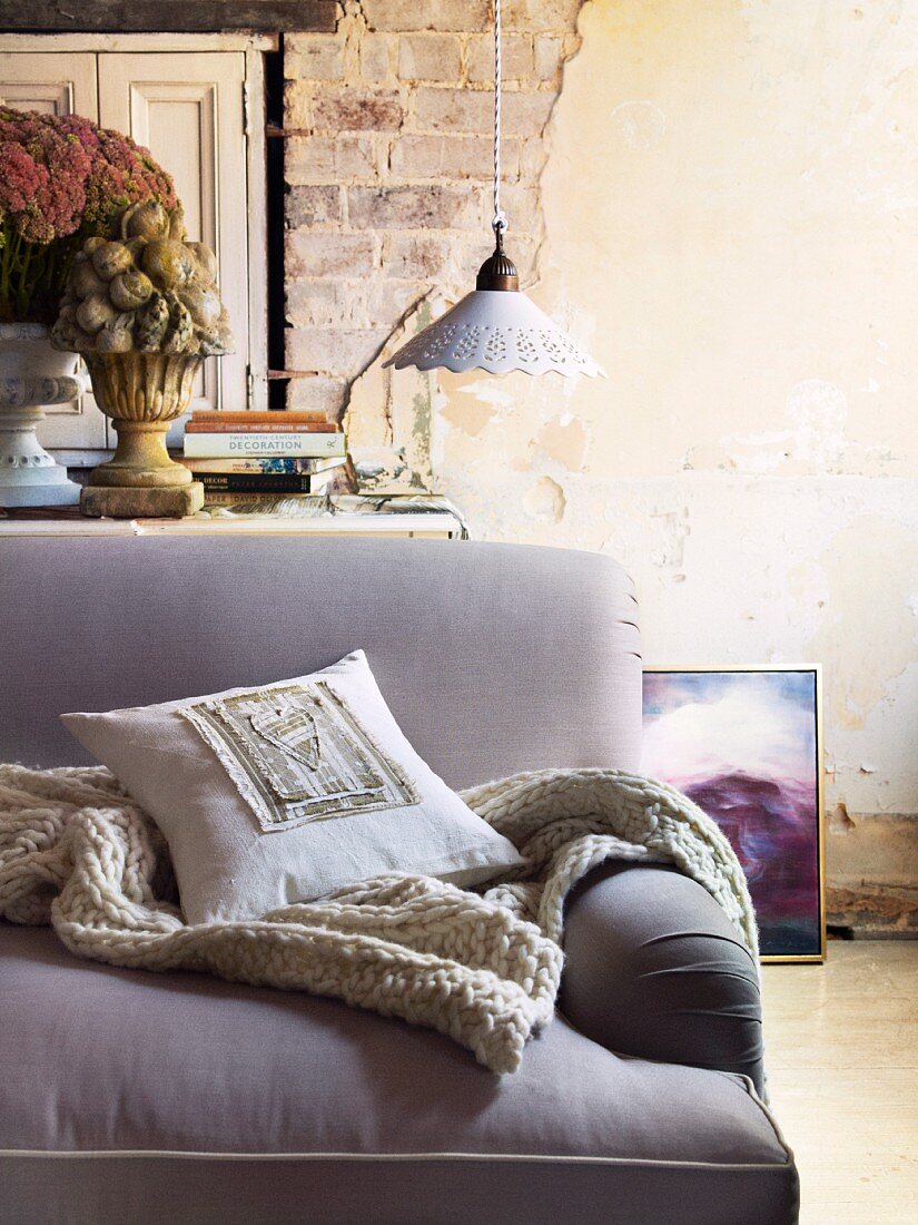 Cushion and knitted blanket on sofa with lilac upholstery in front of vintage pendant lamp and exposed brickwork of wall with crumbling plaster and peeling paint