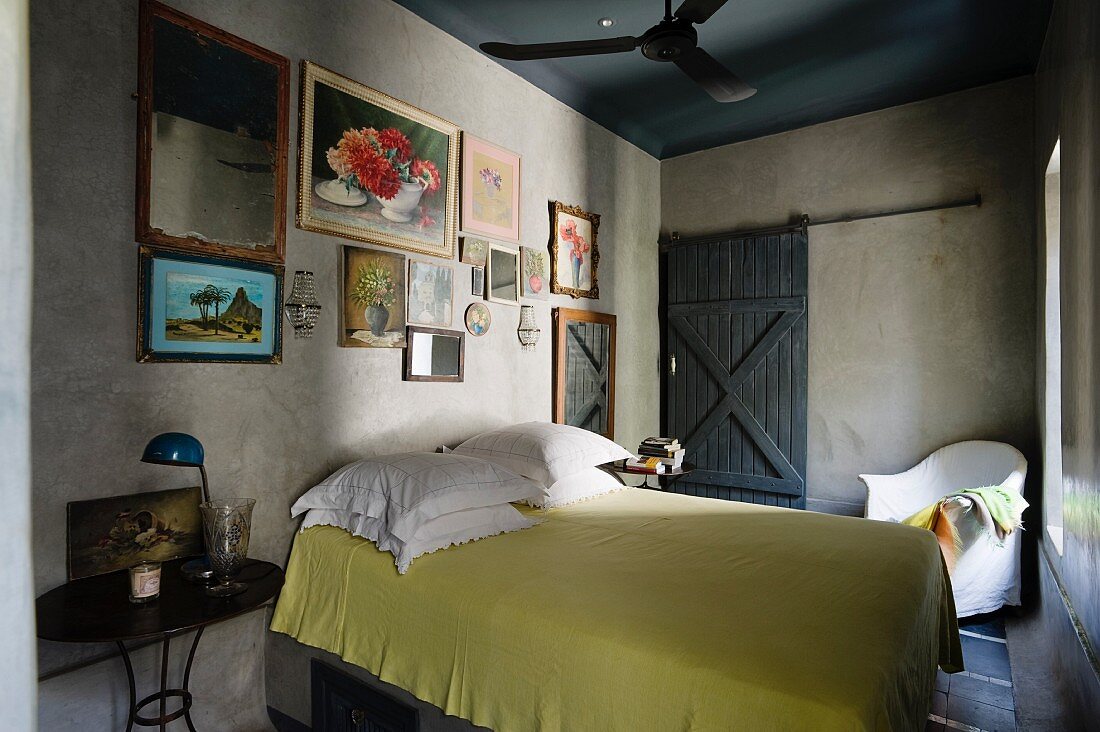 Schlichtes Schlafzimmer mit hölzerner Schiebetür, einer Bildersammlung mit Blumenmotiven an der Wand und einem zitronengelben Bettüberwurf