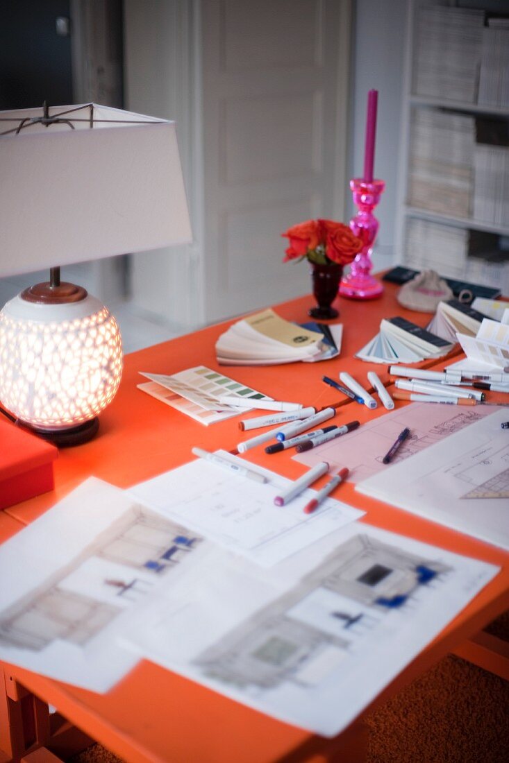 Orangefarbener Arbeitstisch mit Schreib- und Zeichenutensilien; am Tischrand eine leuchtende Tischlampe, Rosen und ein pinkfarbener Kerzenhalter