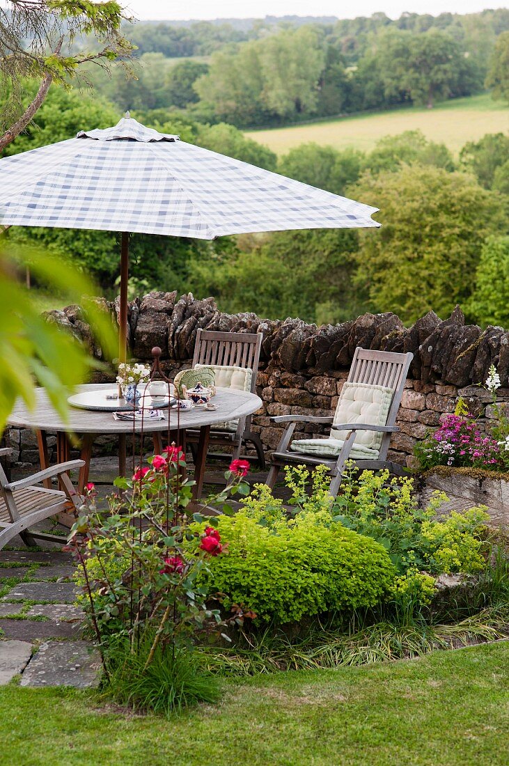 Idyllischer Terrassenplatz in der Gartenecke mit Sitzgruppe und blau-weiß kariertem Sonnenschirm vor Gartenmauer aus Naturstein, im Hintergrund grüne, englische Hügellandschaft