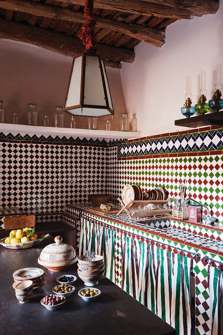 Orientalische Küchenecke mit Mosaik gefliestem Spültisch und eben solchen Wänden; mit Öllampen und Glasgefäßen bestückte Wandboarde bilden den Abschluss zur einfach gestrichenen Wand, im Vordergrund ein Holztisch mit gefüllten Schälchen