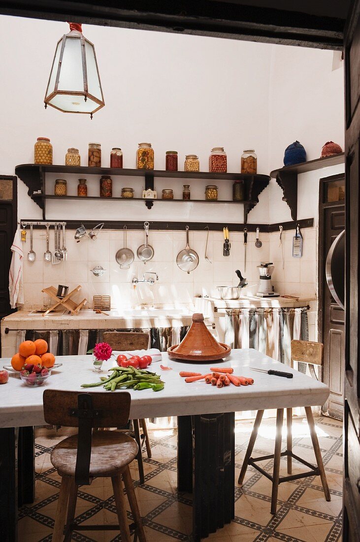 View into simple Oriental kitchen with massive stone table and high wooden stools; chunky sink unit, kitchen utensils hanging from wall rack and jars of preserves on bracket shelf in background