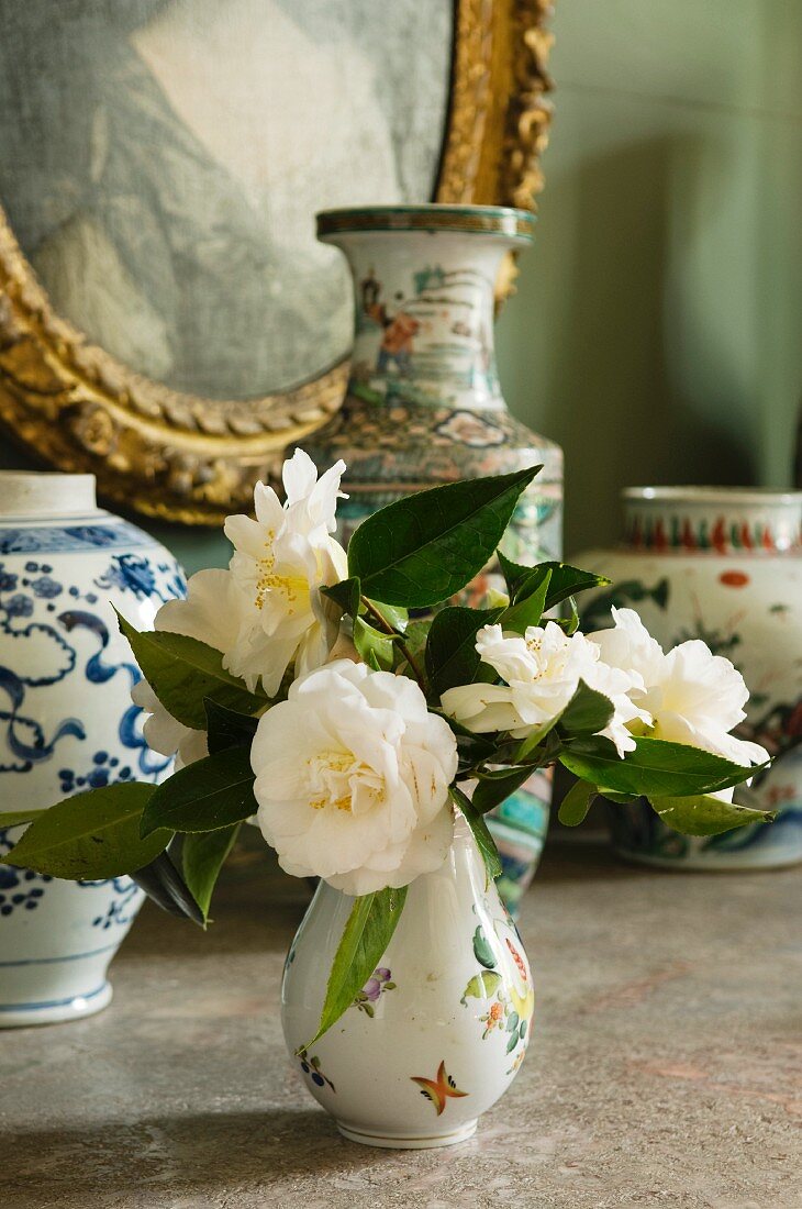 Posy of white roses in china vase in front of collection of vases and gilt-framed picture