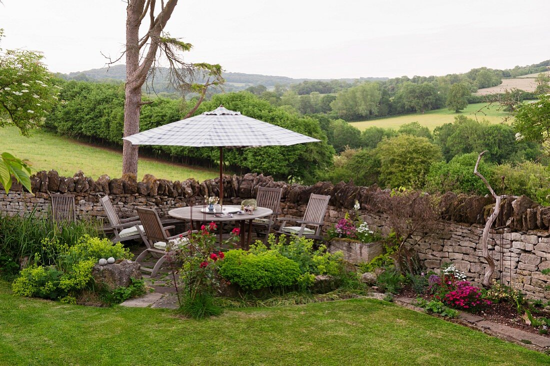 Idyllischer Terrassenplatz in der Gartenecke mit Sitzgruppe und Sonnenschirm vor Gartenmauer aus Naturstein, im Hintergrund grüne, englische Hügellandschaft