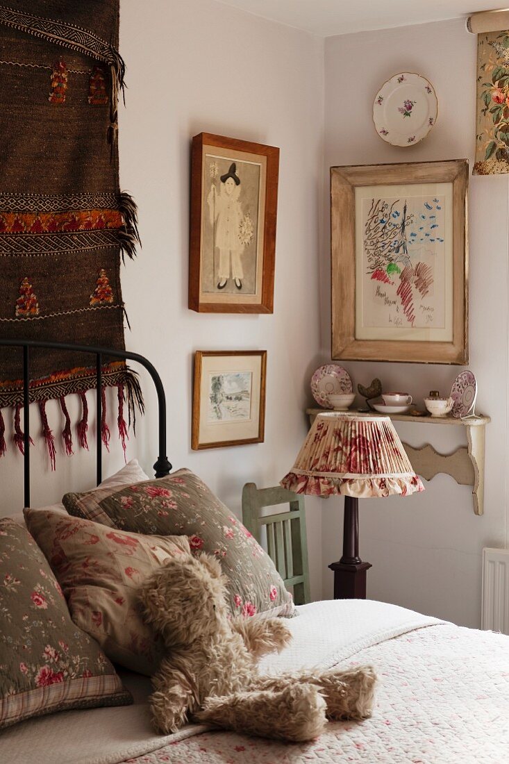 Feminine Schlafzimmerecke mit gerahmten Bildern und einer Wandkonsole mit dekoriertem Porzellan, davor nostalgische Nachtischlampe auf Holzstuhl; kuschliger Teddybär wartet auf dem gemütlichen Bett