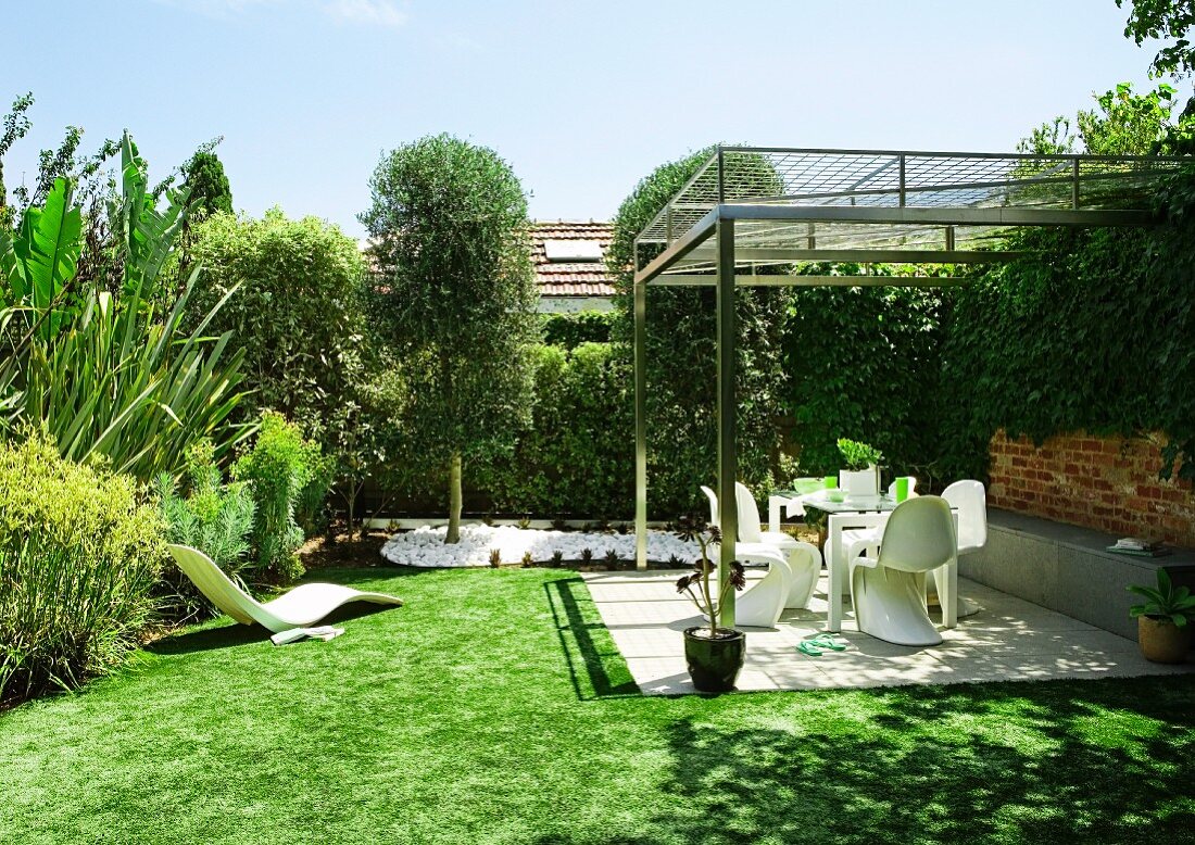 Garten mit gepflasterter Terrasse und lichter Überdachung; im Hintergrund hohe Hecken und verschiedene Gartenpflanzen