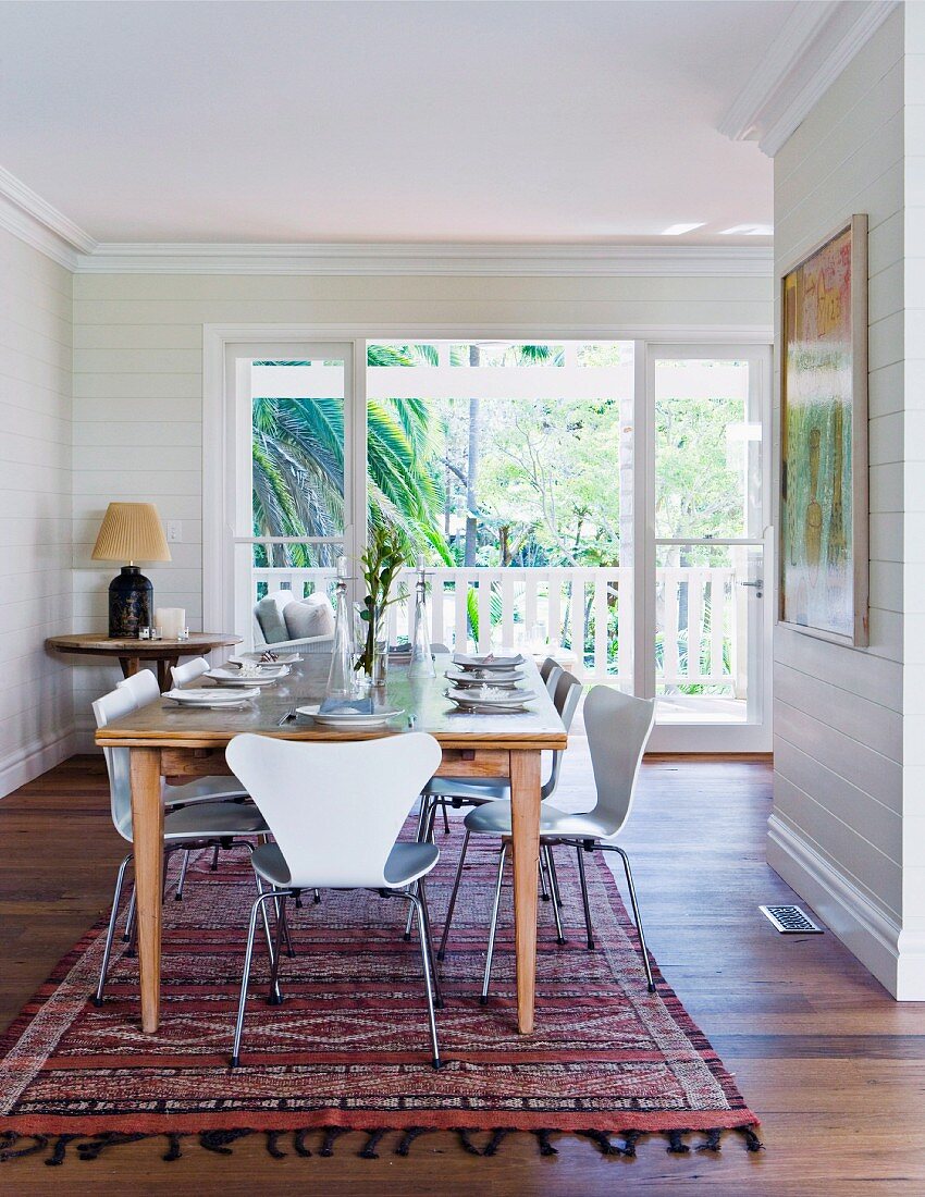 Luftiger Essbereich mit großem Holztisch und weissen Stühlen; im Hintergrund ein sonniger Balkon mit Blick auf eine große Palme