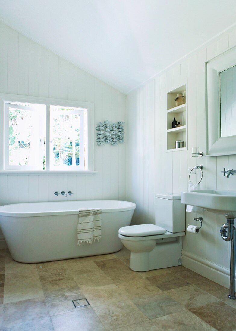 Bright, spacious bathroom under the sloping roof with modern bathtub, stone tiles and large window