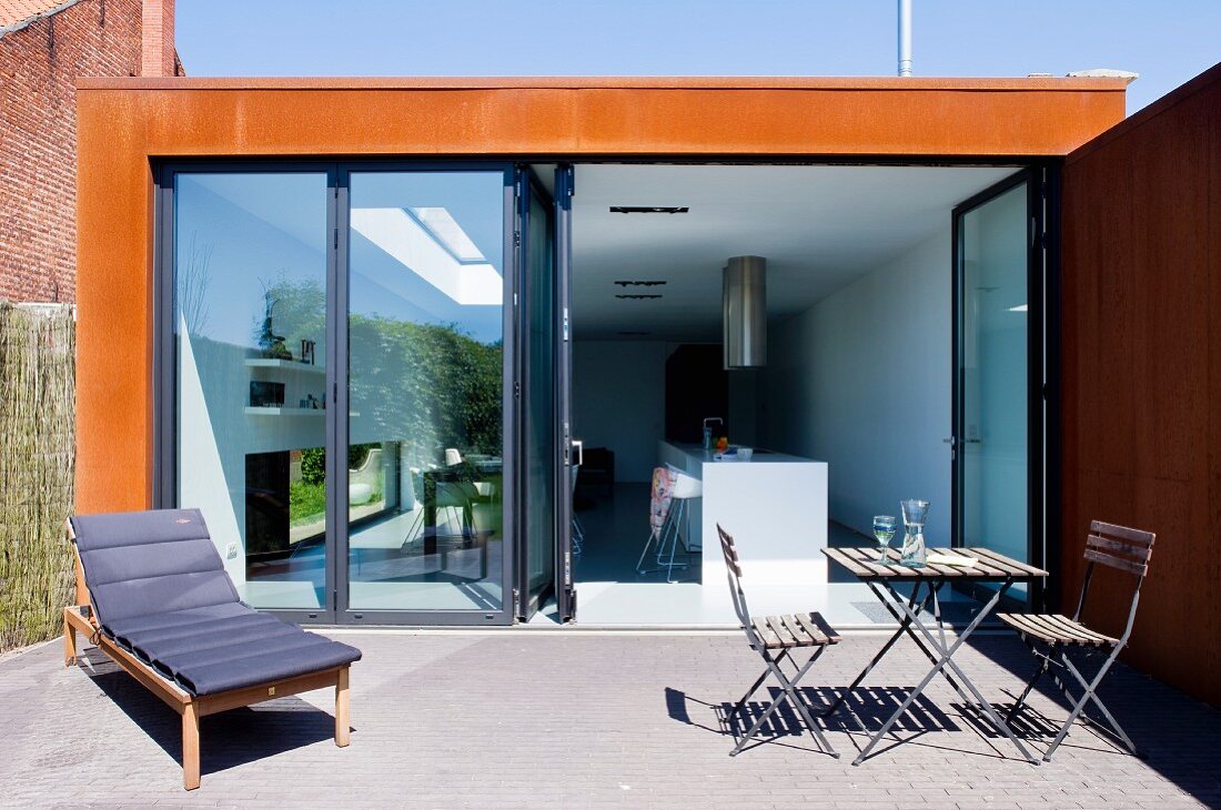 Terrasse mit Liege, Tisch und Stühlen und Blick durch geöffnete Falttür in die Küche