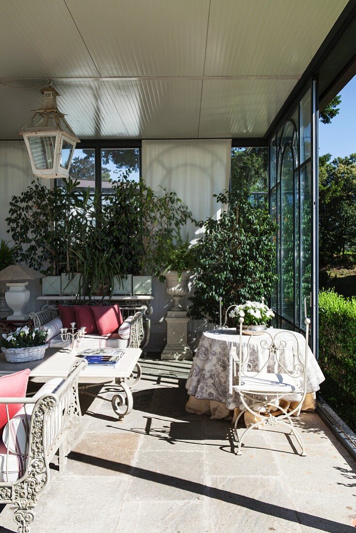 Conservatory with open, sliding glass wall, ornate metal furniture, luxuriant greenery and view of park