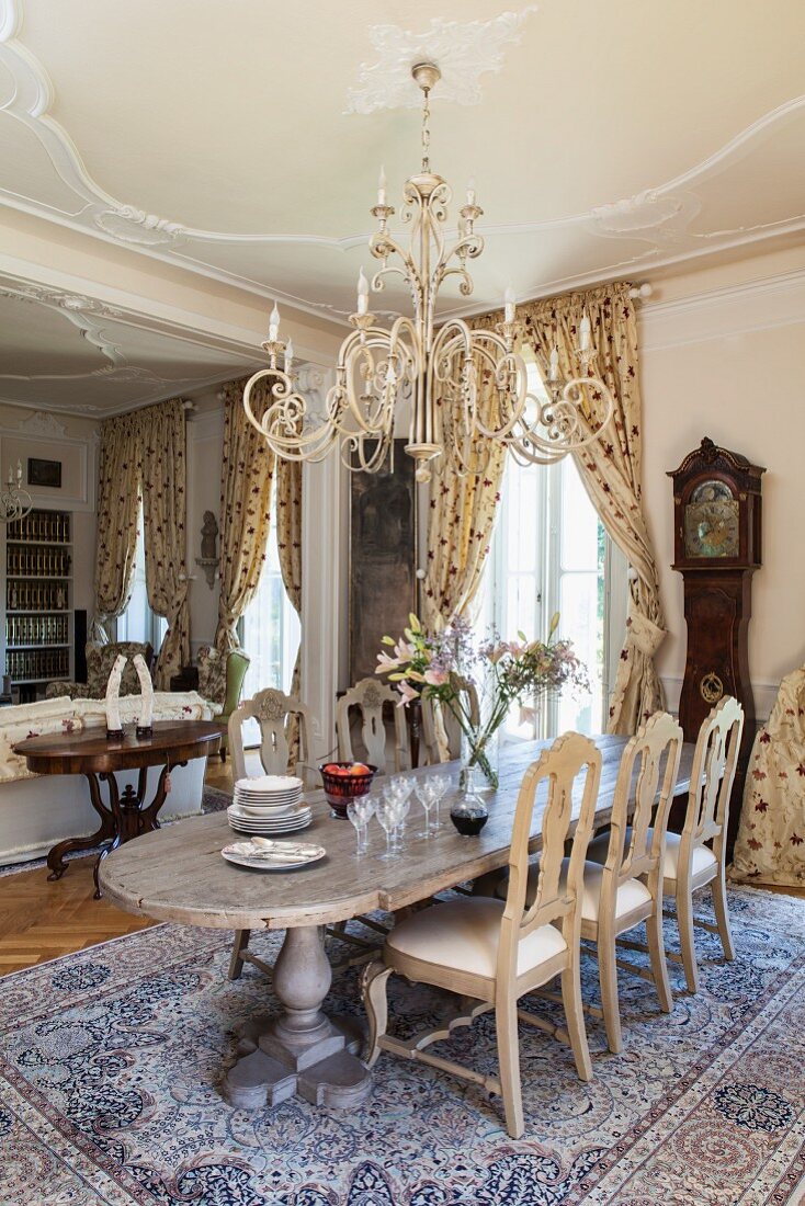 View from dining area with long dining table, fine silk rug and pretty chandelier hanging from stucco ceiling to living area with library