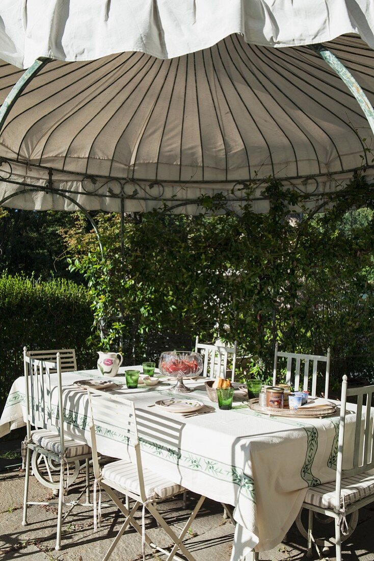 Gedeckter Tisch mit filigranen Metallstühlen unter rundem Dach eines bewachsenen Gartenpavillons