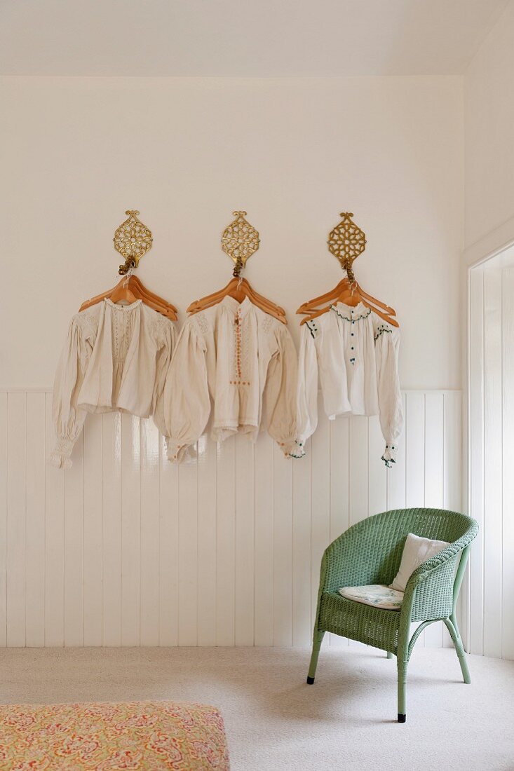 An Haken hängende Kleiderbügel mit ungarischen Trachtenblusen an weisser Wand; darunter ein pastellgrüner Rattanstuhl