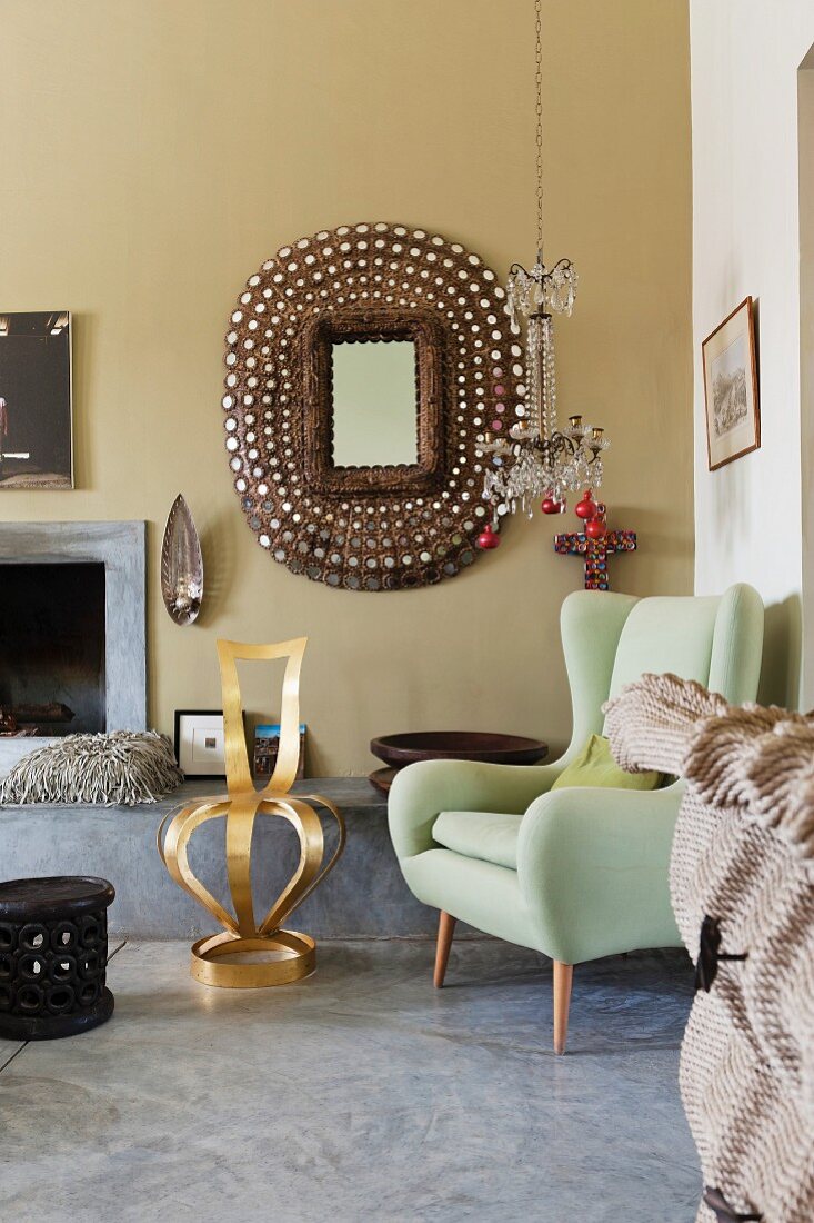 Lindgrüner Ohrensessel im Retrostil und goldfarbener Stuhl vor kunsthandwerklichem Wandspiegel und offenem Kamin