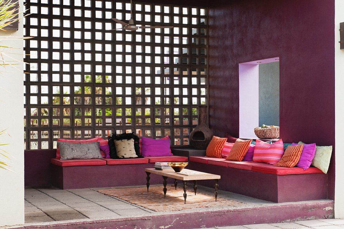 Südamerikanischer Terrassenbereich mit gepolsterter Sitzbank, vielen Kissen und Einblick in den Garten durch eine Holzgitterwand