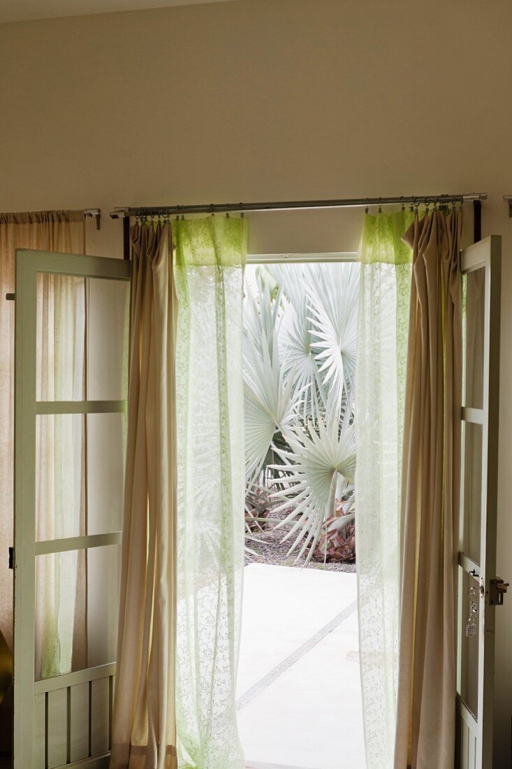 Opened French window with gauzy curtains leading to inviting exotic garden beyond