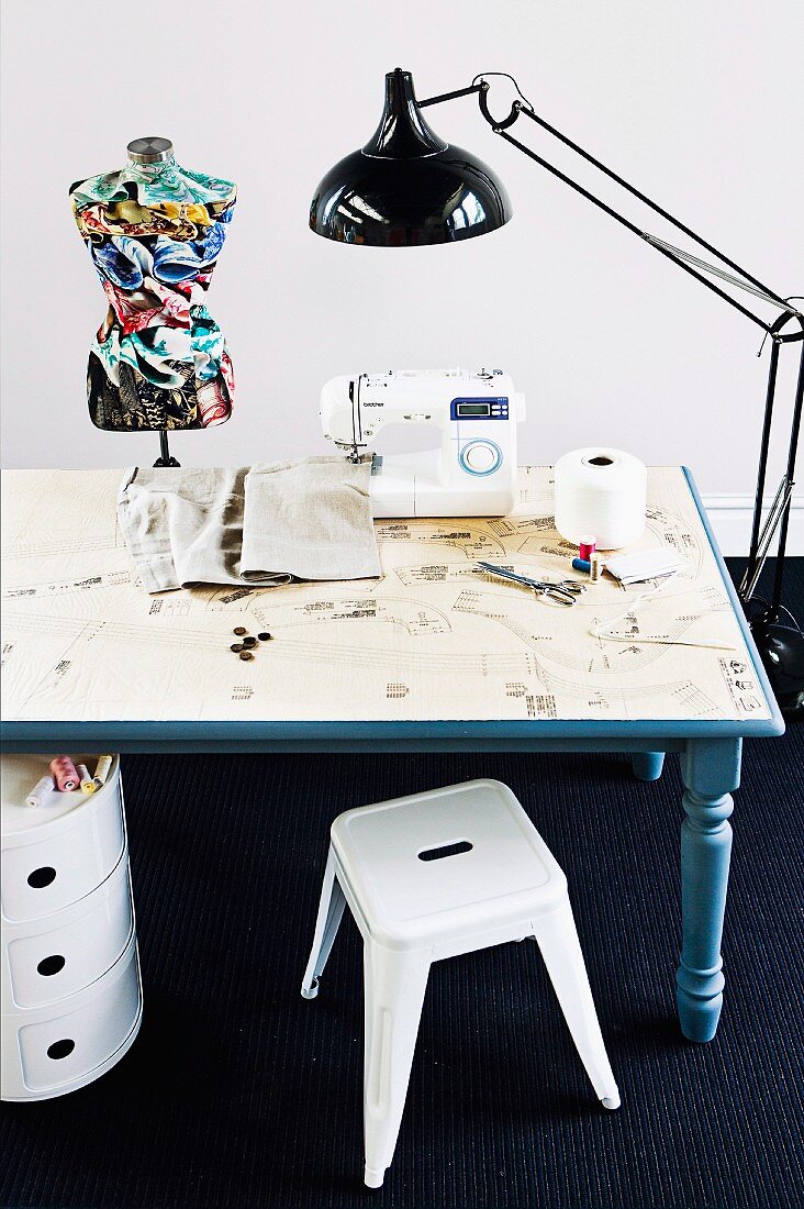Selbstrenovierter, blauer Holztisch mit eingeklebter Schnittmuster-Oberfläche; darauf eine Nähmaschine und eine bunte Kleiderpuppe