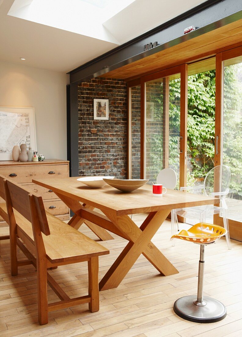 Contemporary, camping-style wooden table and bench in front of brick wall and large windows