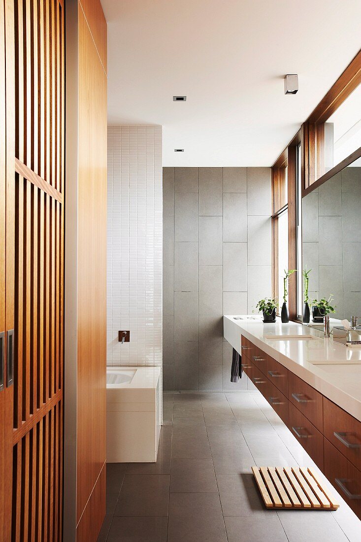 Designerbad - Zwischen Wänden eingespannter Waschtisch mit weisser Platte in grau gefliestem Bad und Einbauschrank mit Holzlamellentür