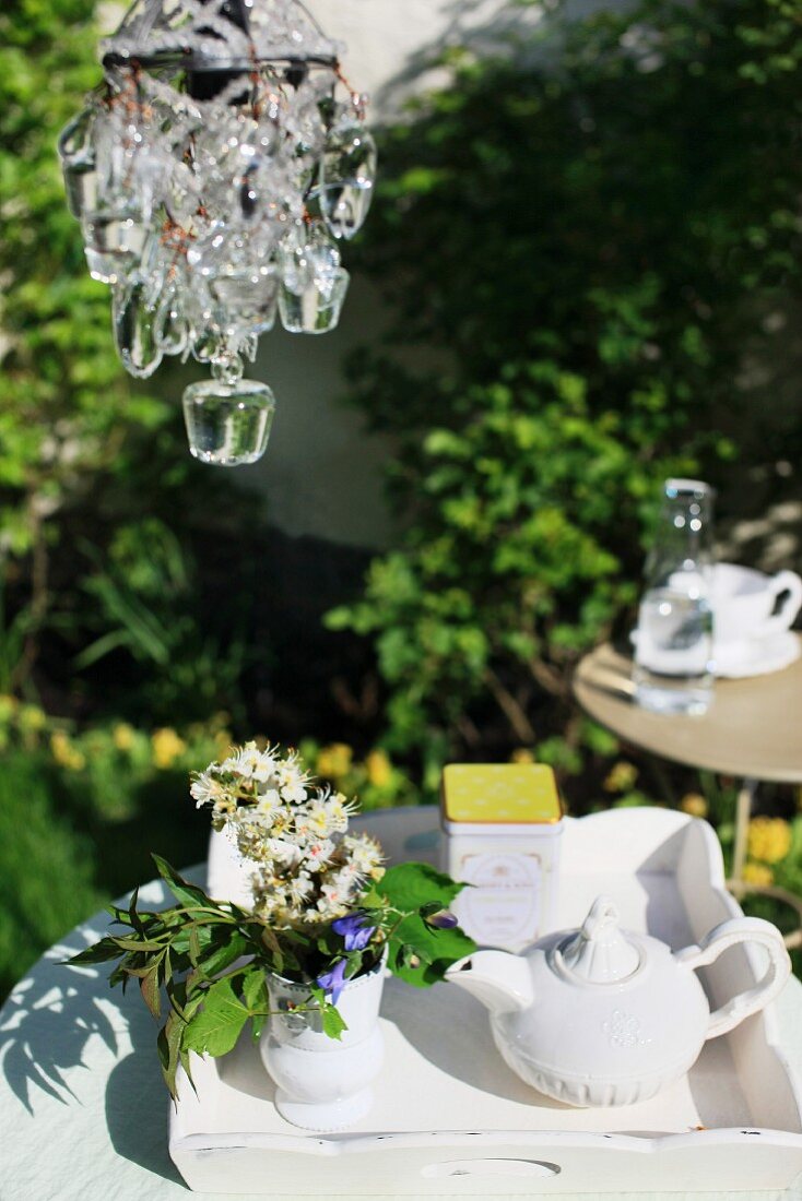 Kronleuchter über einem Tablett mit Porzellankanne, Teedose und Tischblumen im sonnigen Garten