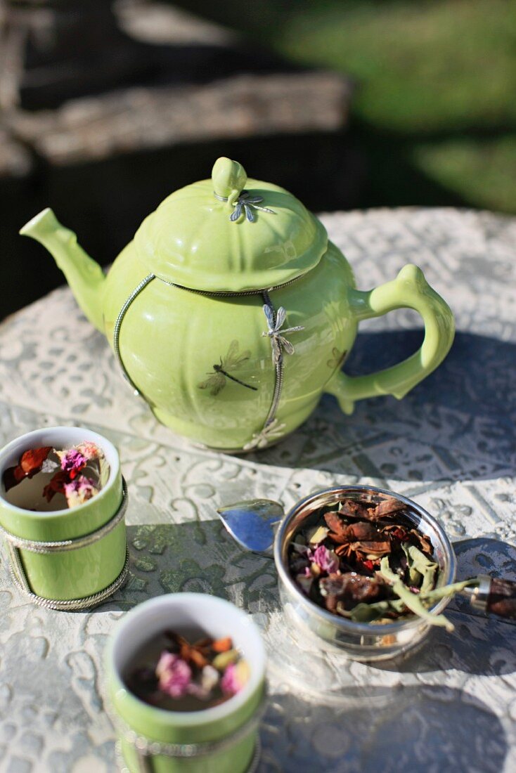 Lindgrüne Teekanne mit silbernem Libellenmotiv und passende Teebecher auf einem antiken Gartentisch