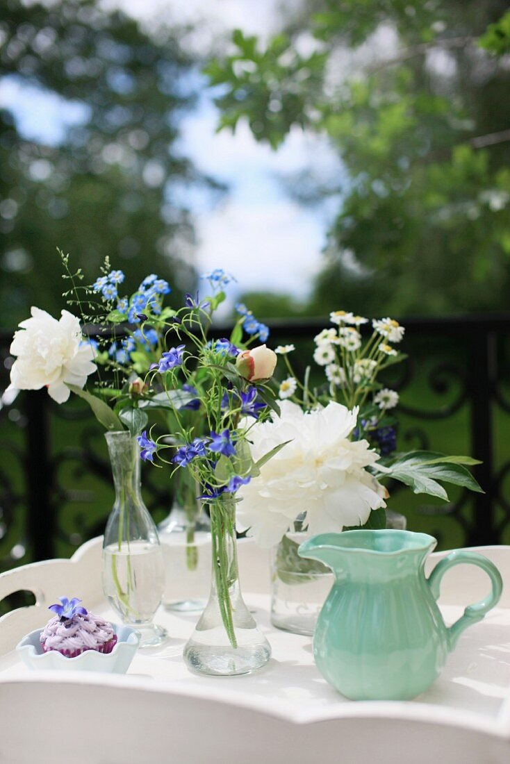 Kleine Glasvasen mit duftigen Sommerblumen und türkise Porzellankanne auf weißem Holztablett