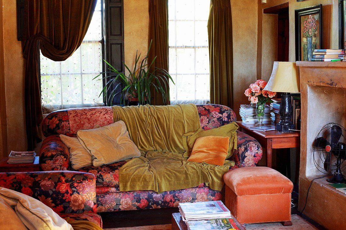 Sofa mit Blumenmuster und Uni-Samt als vorherrschende Dekostoffe in dicht möbliertem Vintage Wohnzimmer mit offenem Kamin