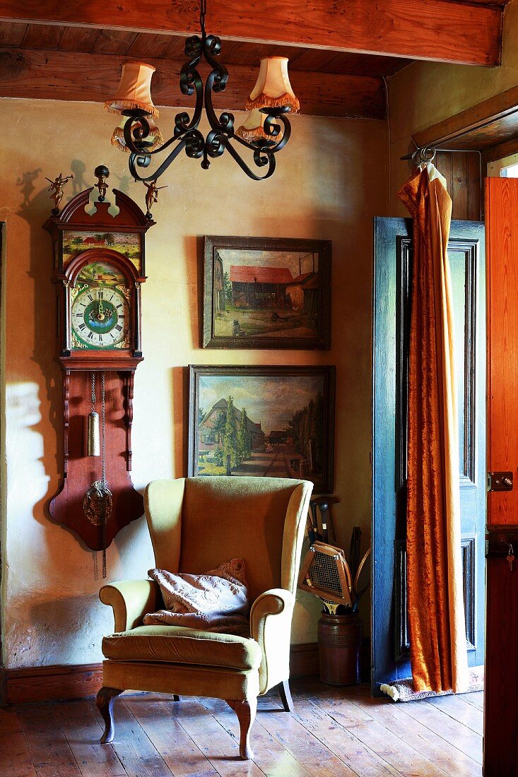 Wing-back chair and wrought iron chandelier in front of antique wall clock and old landscape paintings
