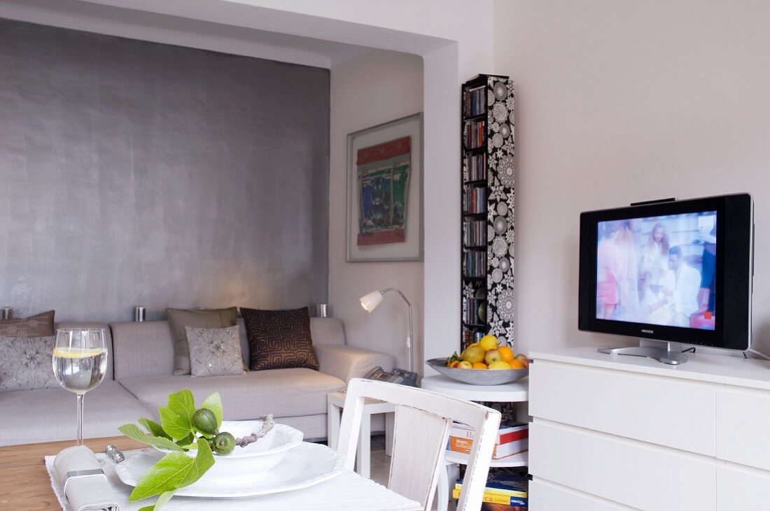 Alkoven mit weisser Wohnzimmercouch vor grauer Wand; davor ein gedeckter Esstisch und ein laufendes Fernsehgerät