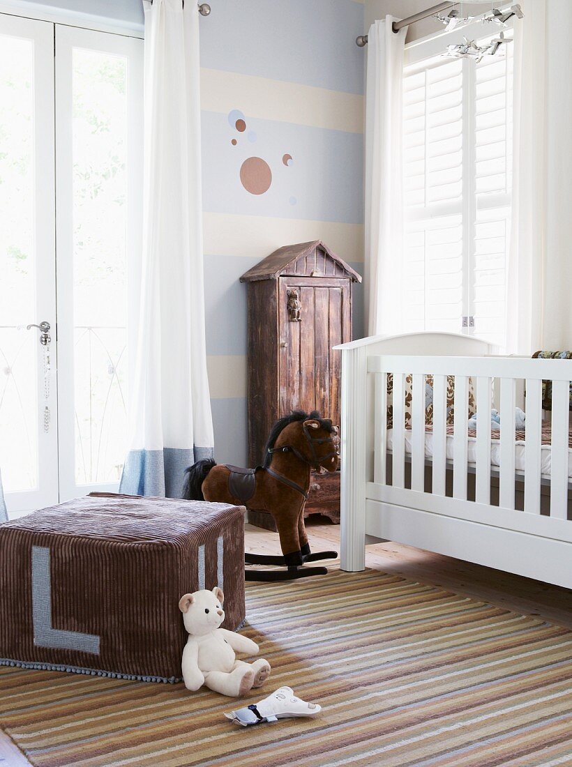 Nostalgisch angehauchtes Kinderzimmer mit weißem Gitterbettchen, Schaukelpferd und großem Sitzwürfel aus Velour; an der blau-weiß gestrichenen Wand steht ein Holzschränkchen mit Dächchen