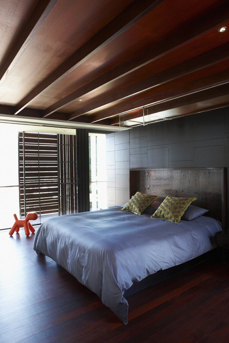 Schlafzimmer mit edler Holzdecke und sonnengeschützter Glasfront durch verschiebbare Holzpaneele; in der Mitte ein großes Doppelbett
