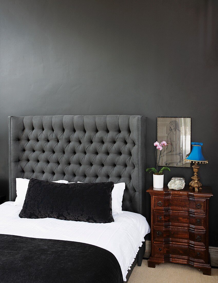 Doppelbett mit gestepptem Kopfende Ton in Ton an anthrazitfarbener Wand mit weisser Bettwäsche und schwarzem Überwurf; daneben ein antikes Nachtkästchen