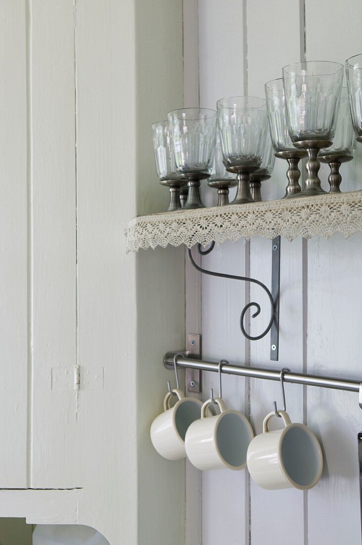 Glasses on wall-mounted shelf with lace trim above cups hanging on rail in corner of white, wood-panelled kitchen