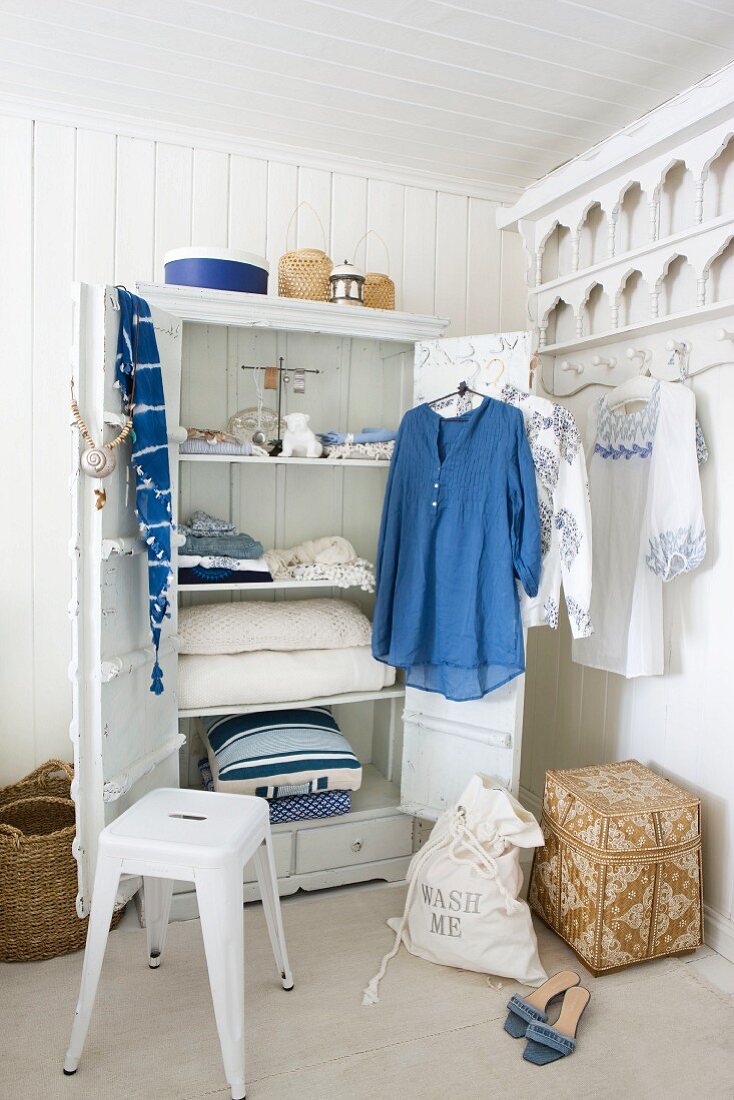 Blue and white items of clothing hanging on door of open wardrobe surrounded by baskets, laundry bag and white metal stool