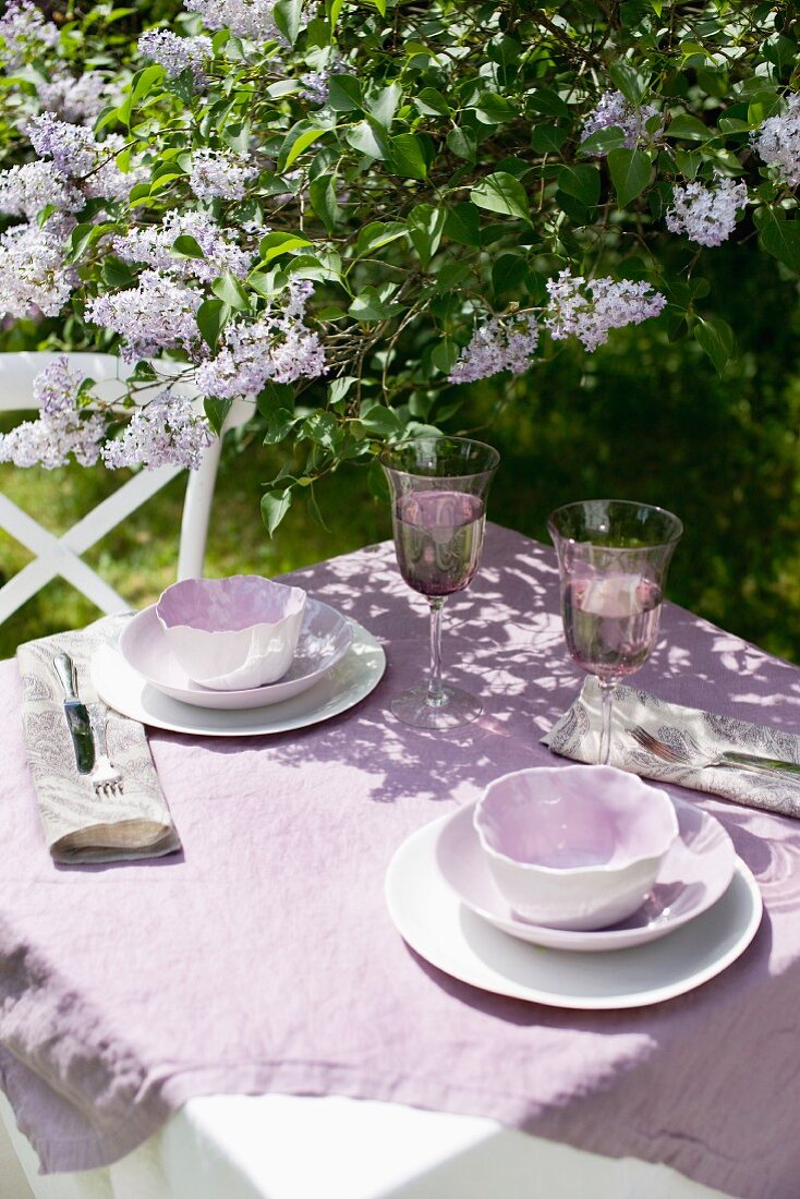 Garden table in sunshine with violet linen tablecloth and china crockery of the same shade below flowering lilac